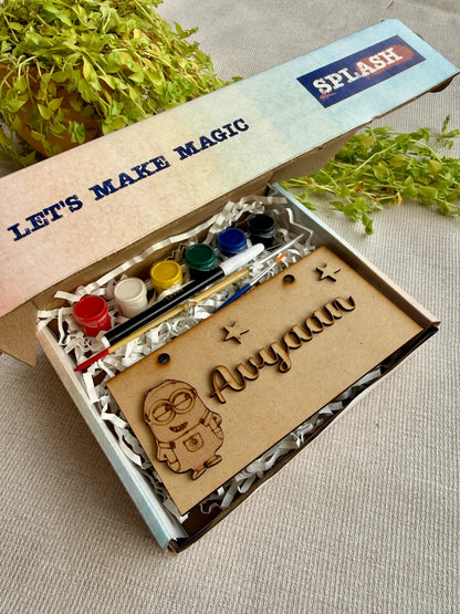 A personalized kids art kit in a cardboard box with a laser-engraved nameplate reading 'Ayyan' and a Minion illustration. The kit includes paint pots, a paintbrush, and a palette, with the box lid showing 'LET'S MAKE MAGIC' and 'SPLASH'.