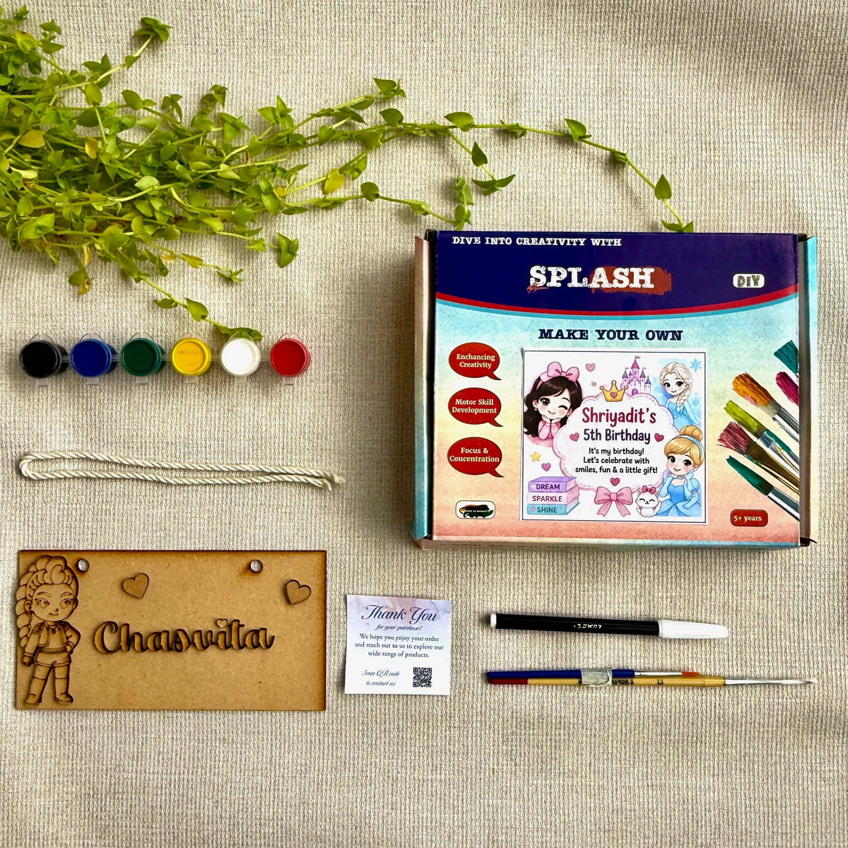 A custom DIY embroidery kit laid out on a beige linen background, including a colorful 'SPLASH' branded box, embroidery floss, a wooden nameplate with an embroidered character, pens, and a small card.