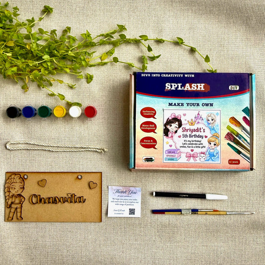 A custom DIY embroidery kit laid out on a beige linen background, including a colorful 'SPLASH' branded box, embroidery floss, a wooden nameplate with an embroidered character, pens, and a small card.