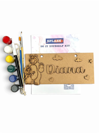A DIY wooden engraving kit labeled 'SPLASH DO IT YOURSELF KIT' on a white background, featuring a natural wood plaque engraved with a bear, clouds, hearts, and the name 'Diana', alongside paint pots, brushes, and a paint palette.