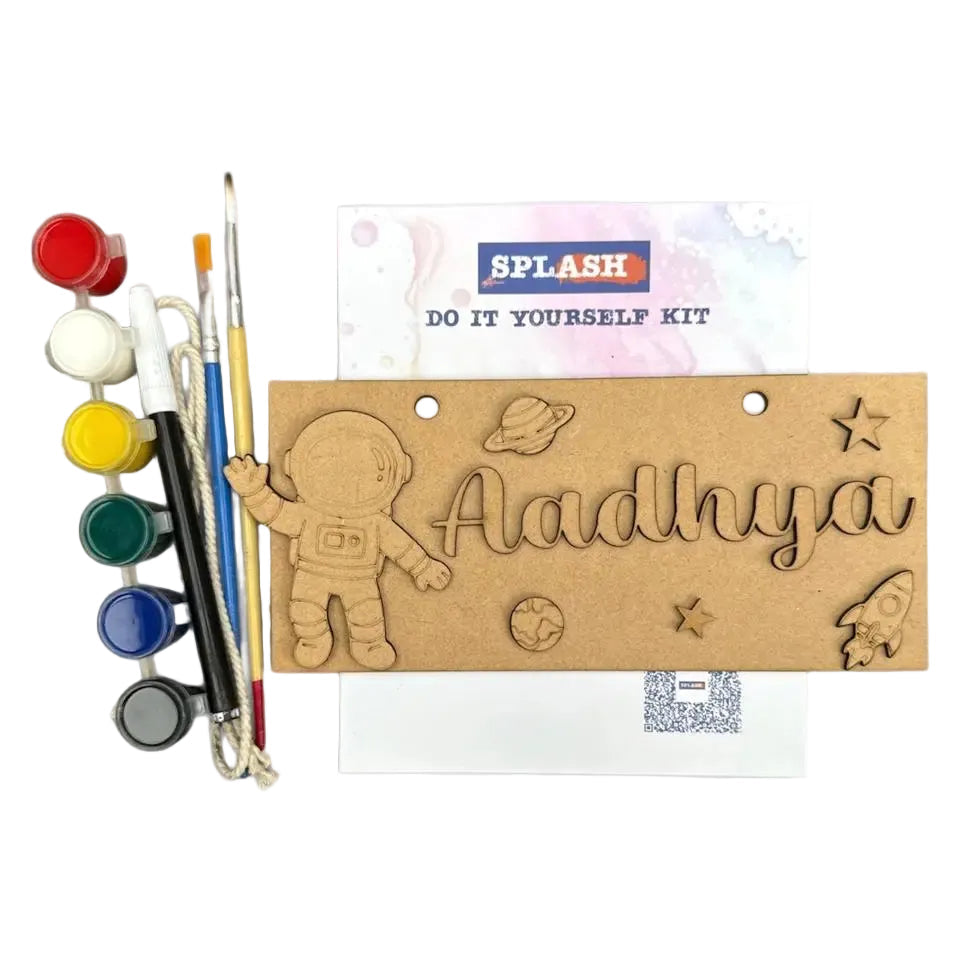 A personalized kids' do-it-yourself painting kit by Splash, featuring a wooden astronaut-themed plaque engraved with the name 'Aadhya' and space motifs, alongside paint pots, paintbrushes, and a card on a white background.
