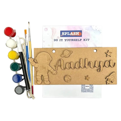 A personalized kids' do-it-yourself painting kit by Splash, featuring a wooden astronaut-themed plaque engraved with the name 'Aadhya' and space motifs, alongside paint pots, paintbrushes, and a card on a white background.