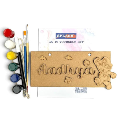 A DIY wooden name carving kit shown on a white background, featuring a natural wood plaque engraved with the name 'Aadhya', a carved teddy bear, and small decorative shapes. The kit includes paint pots in multiple colors, paintbrushes, and a printed instruction card with the brand name 'SPLASH'.