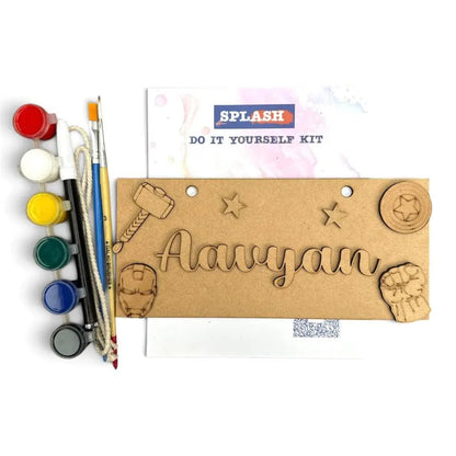 A DIY craft kit labeled 'SPLASH DO IT YOURSELF KIT' on a white background, featuring a brown cardboard plaque engraved with the name 'Aavyan' and small decorative icons, alongside paint pots, brushes, and a palette.