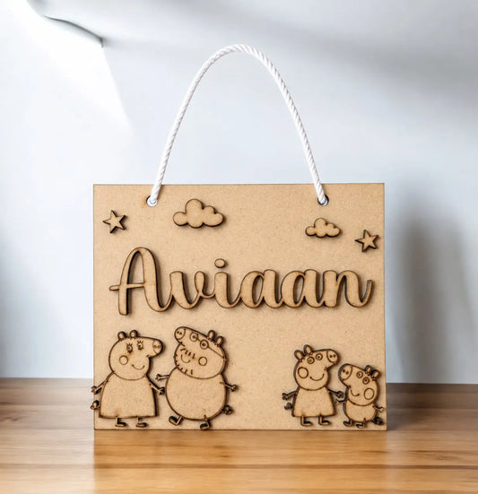 A personalized kraft paper gift bag with a white rope handle, featuring laser-cut engravings of the name 'Aviaan', clouds, stars, and Peppa Pig characters. The bag is displayed on a wooden surface against a white background.
