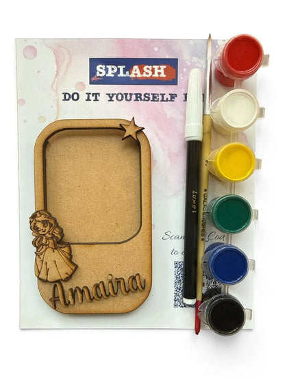 A DIY craft kit featuring a laser-cut wooden frame with a personalized name 'Amaira' and a small wooden doll figure, shown next to a paint palette with six paint wells (red, white, yellow, green, blue, black), a paintbrush, and a marker on a white background.