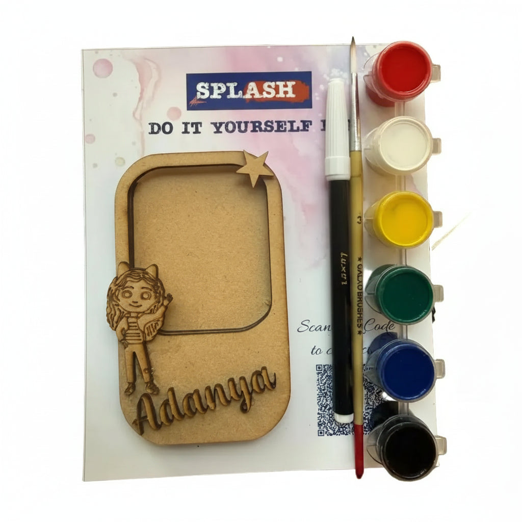 A DIY craft kit labeled SPLASH on a wooden surface, featuring a laser-cut wooden frame with a cartoon character and the name 'Adanya', plus paint pots, a paintbrush, and a marker.