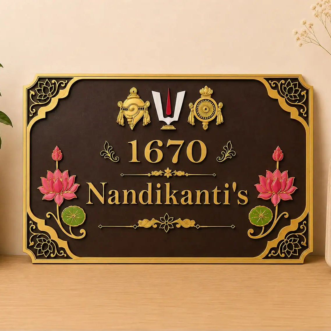A decorative rectangular wall sign with a dark brown background and ornate gold border, featuring the text '1670 Nandikanti's' with pink lotus flowers and gold decorative motifs. The sign is displayed on a light wood surface against a beige wall, flanked by a potted plant on the left and a vase with dried stems on the right.