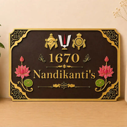 A decorative rectangular wall sign with a dark brown background and ornate gold border, featuring the text '1670 Nandikanti's' with pink lotus flowers and gold decorative motifs. The sign is displayed on a light wood surface against a beige wall, flanked by a potted plant on the left and a vase with dried stems on the right.