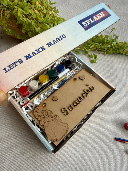 A personalized kids' craft kit in an open box, featuring a wooden name plaque engraved with 'Gaanchi' and a fairy illustration, alongside small paint pots, a paintbrush, and decorative paper on a neutral fabric background.