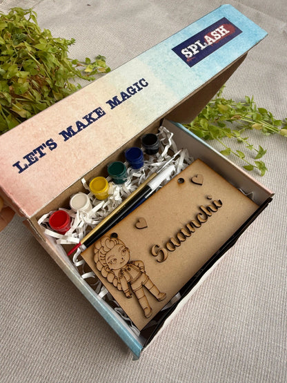 A personalized kids' craft kit in an open cardboard box, featuring acrylic paint pots, a paintbrush, and a wooden nameplate engraved with 'Saaachi' and a fairy illustration. The box lid has 'LET'S MAKE MAGIC' and 'SPLASH' printed on it, set on a neutral fabric background with green foliage.