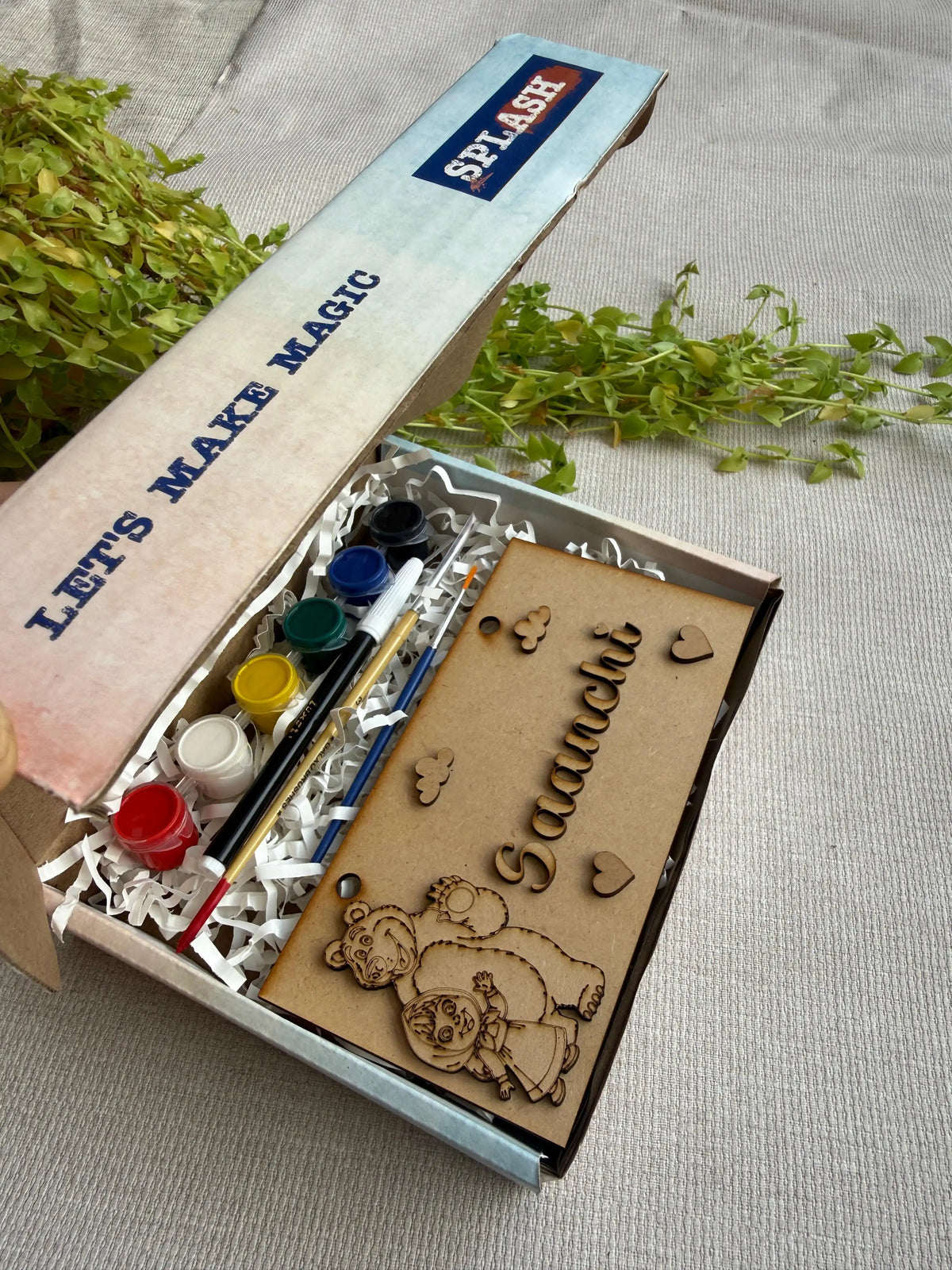 A boxed craft kit opened on a light fabric surface with greenery props. The kit includes a wooden bear-shaped plaque with laser-cut text and heart motifs, paint tubes, brushes, and a box printed with 'LET'S MAKE MAGIC' and 'SPLASH'.