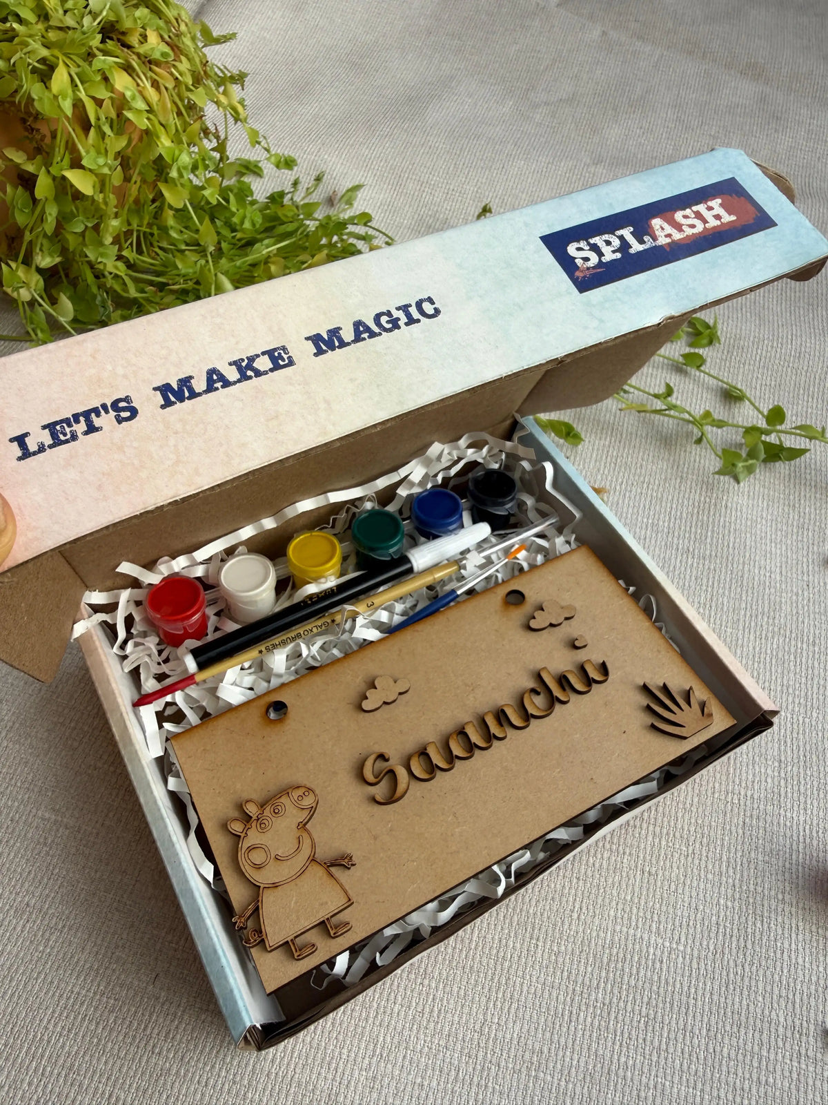 A personalized kids' paint kit in an open cardboard box with a lid reading 'LET'S MAKE MAGIC' and 'SPLASH'. The kit includes acrylic paint cups, a paintbrush, and a wooden nameplate engraved with 'Gaanchu' and a cartoon character, set on a neutral fabric background with green foliage.