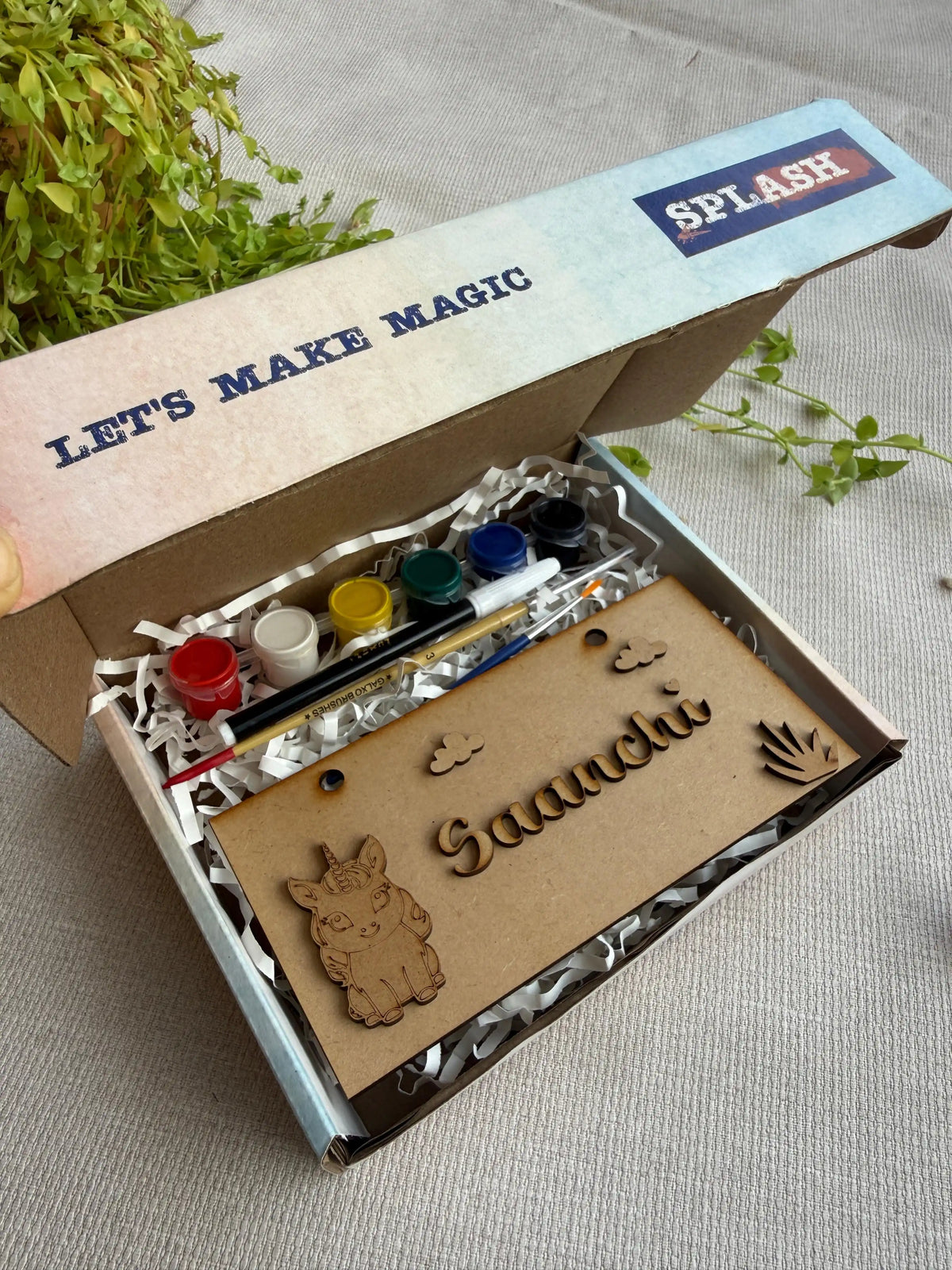 A children's craft kit in an open cardboard box with the brand name SPLASH visible on the lid. The kit includes small paint bottles, a paintbrush, and a wooden nameplate engraved with 'Saaachi' and decorative animal and hand motifs, set on a neutral fabric surface with green leaves in the background.