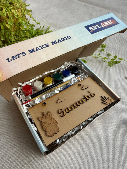 A children's craft kit in an open cardboard box with the brand name SPLASH visible on the lid. The kit includes small paint bottles, a paintbrush, and a wooden nameplate engraved with 'Saaachi' and decorative animal and hand motifs, set on a neutral fabric surface with green leaves in the background.