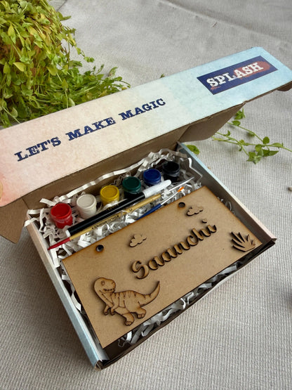 A personalized children's craft kit in an open cardboard box, featuring a wooden dinosaur nameplate engraved with 'Saanachi' and a handprint, alongside small paint bottles and a paintbrush. The box lid is printed with 'LET'S MAKE MAGIC' and 'SPLASH'.