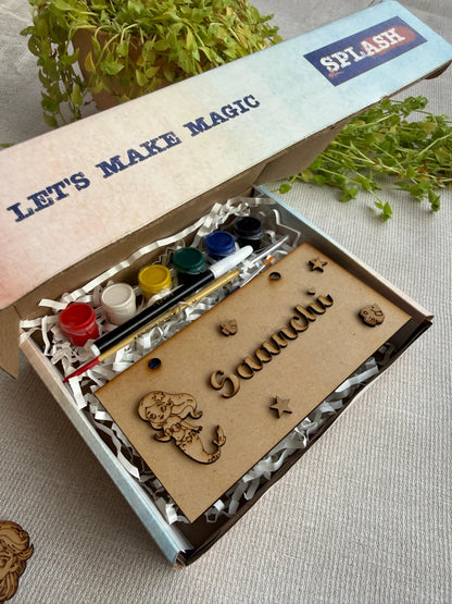 A personalized children's painting kit in a gift box, featuring a wooden nameplate engraved with 'Gaaachi', wooden animal and star cutouts, a paint palette with small paint pots, and a paintbrush. The open box lid displays the text 'LET'S MAKE MAGIC' and 'SPLASH'.