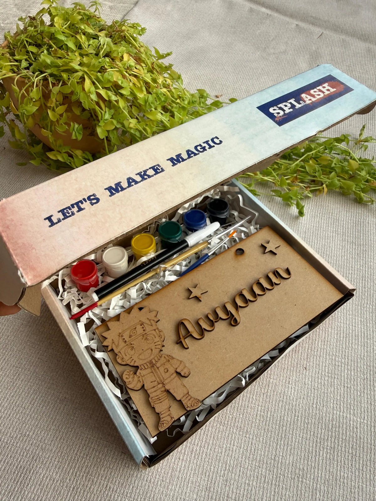 A personalized children's craft kit in an open box, featuring a wooden cutout of a cartoon character and the name 'Aryan' in laser-cut letters, alongside small paint tubes and a paintbrush. The box lid is printed with 'LET'S MAKE MAGIC' and 'SPLASH'.
