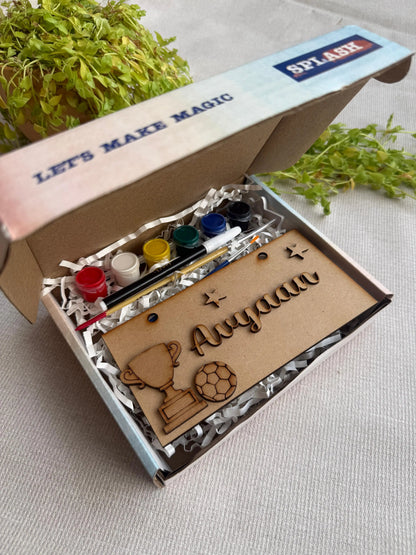 A personalized kids' paint painting kit in a cardboard box, featuring a wooden nameplate engraved with 'Anyaan', a trophy, a soccer ball, and paint bottles, set on a light fabric surface with green plants in the background.