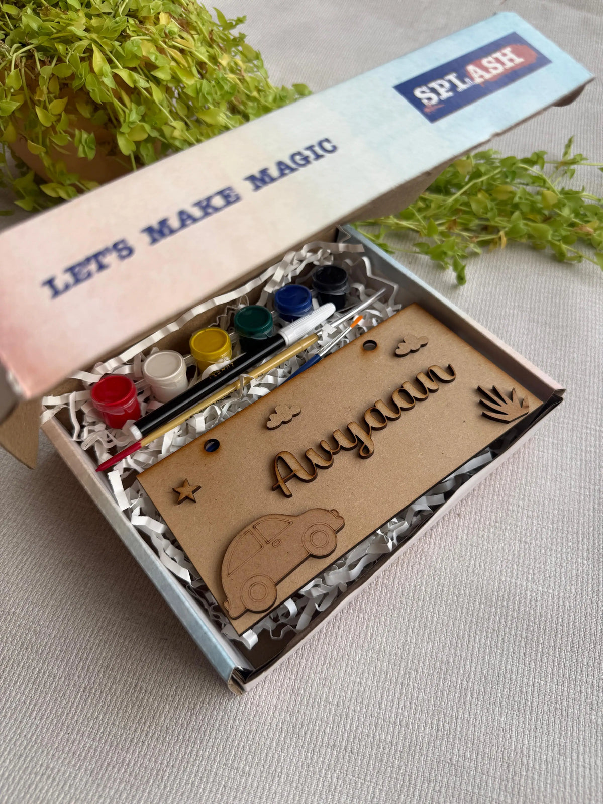 A personalized kids' art kit in a light blue box with the brand name SPLASH. The kit contains acrylic paint pots, a paintbrush, and a wooden nameplate engraved with 'Aaryaan' and decorated with a car and sun shapes, displayed on a neutral fabric background with green plants.