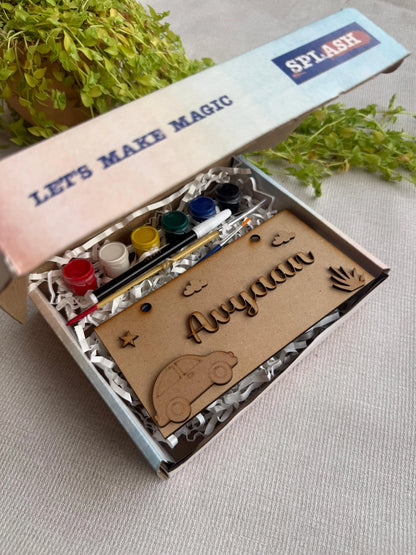 A personalized kids' art kit in a light blue box with the brand name SPLASH. The kit contains acrylic paint pots, a paintbrush, and a wooden nameplate engraved with 'Aaryaan' and decorated with a car and sun shapes, displayed on a neutral fabric background with green plants.