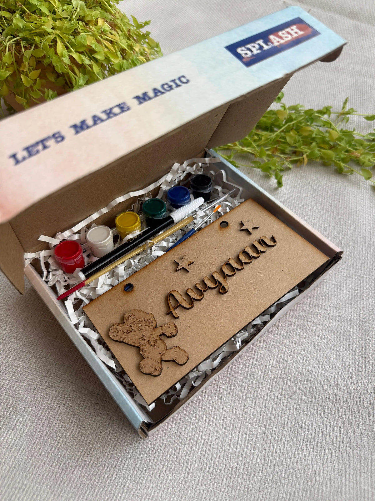 A personalized kids' craft kit in an open cardboard box, featuring acrylic paint bottles, a paintbrush, a wooden bear cutout, and a custom engraved name card reading 'Ayaan'. The box lid has 'SPLASH' printed on it, and the kit is styled with green leaves on a neutral fabric background.