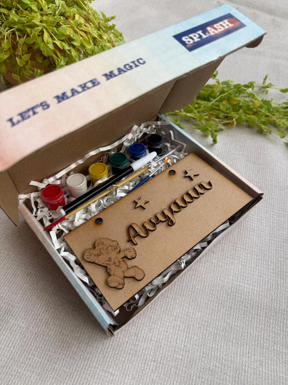 A personalized kids' craft kit in an open cardboard box, featuring acrylic paint bottles, a paintbrush, a wooden bear cutout, and a custom engraved name card reading 'Ayaan'. The box lid has 'SPLASH' printed on it, and the kit is styled with green leaves on a neutral fabric background.
