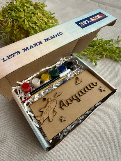 A personalized kids' craft kit in an open cardboard box, featuring wooden laser-cut pieces, a small airplane cutout, and a nameplate reading 'Aloyna', alongside small paint pots and brushes. The box lid has 'SPLASH' branding and 'LET'S MAKE MAGIC' printed on it, with green foliage in the background.