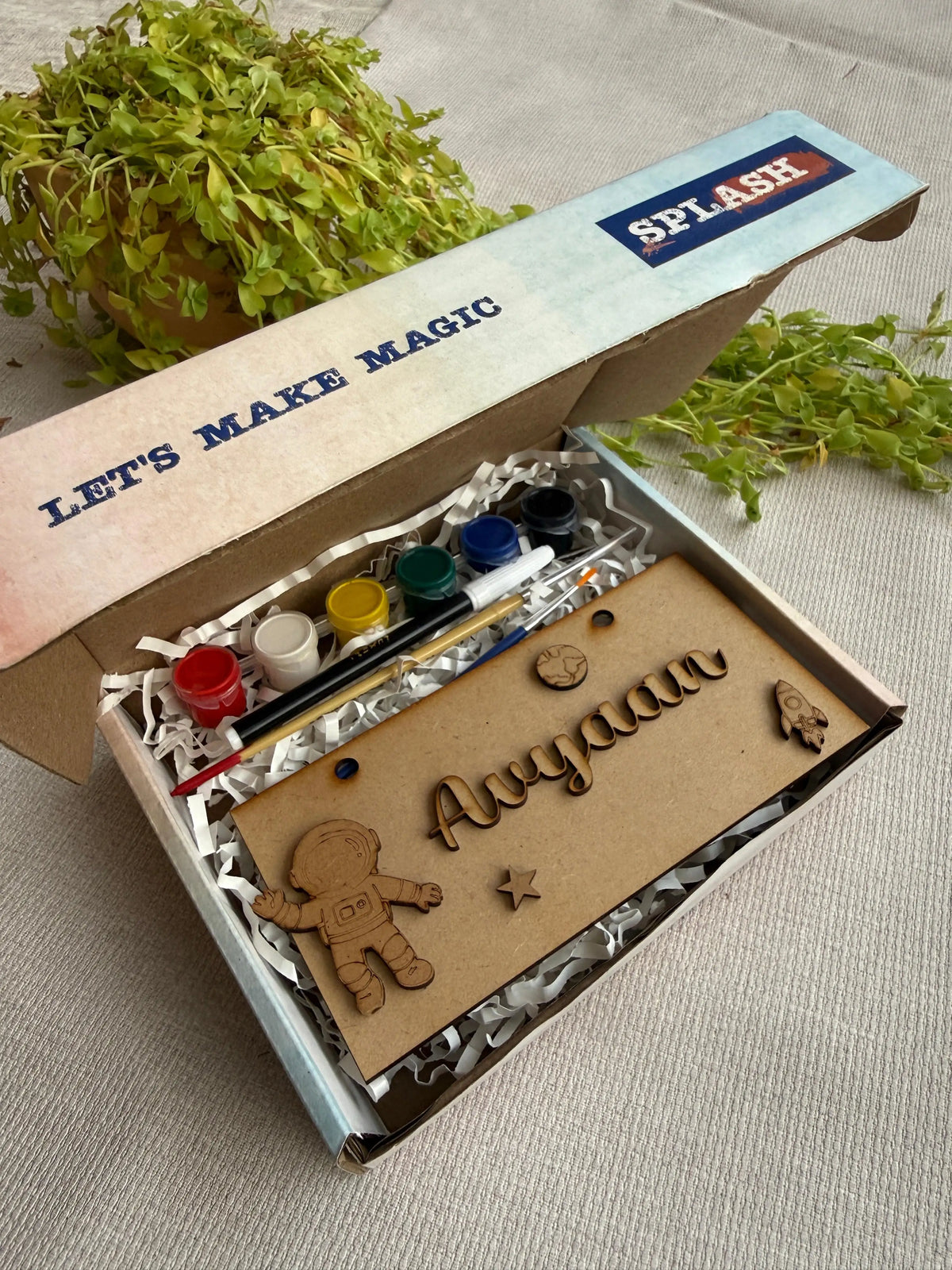 A personalized kids' craft kit in a cardboard box, featuring a wooden astronaut nameplate engraved with 'Ayyan', paint pots, a paintbrush, and laser-cut wooden space-themed decorations. The box lid has 'LET'S MAKE MAGIC' and 'SPLASH' printed on it, and the box is styled with green plants on a neutral fabric background.