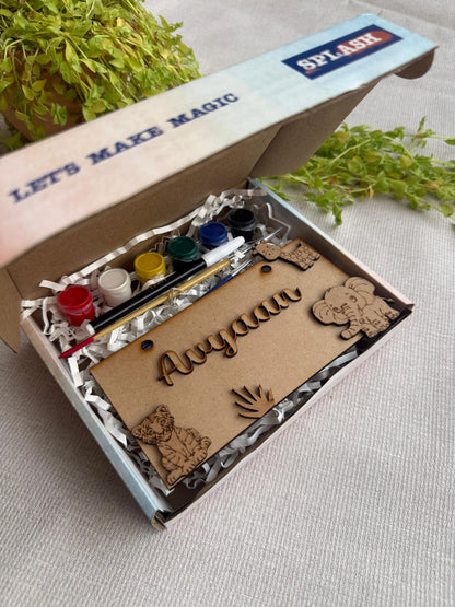 A personalized wooden name art kit in a branded box, featuring laser-cut wooden names and animal shapes, a small paint palette, and a paintbrush on a textured light beige fabric background with green plants.