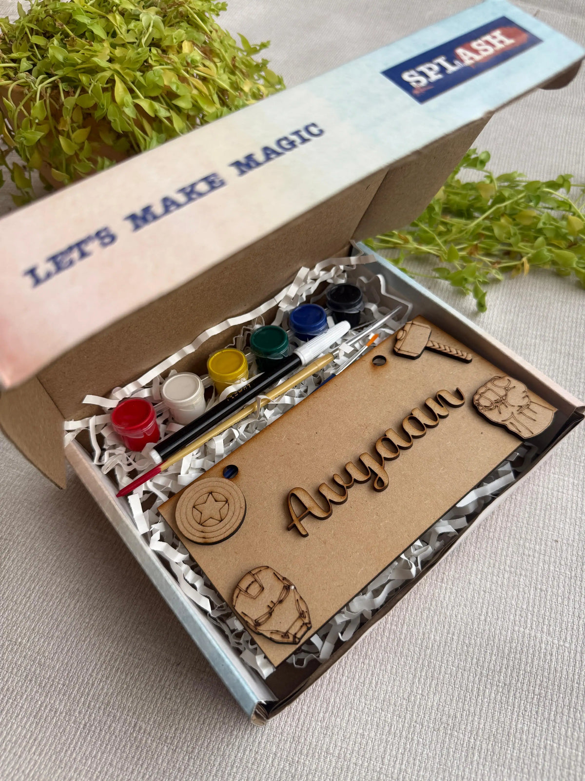 A personalized kids' craft kit in an open cardboard box with the name 'Anyaan' laser-engraved on a brown card. The kit includes small paint pots, a paintbrush, and wooden craft pieces, displayed on white paper shred with green foliage in the background.