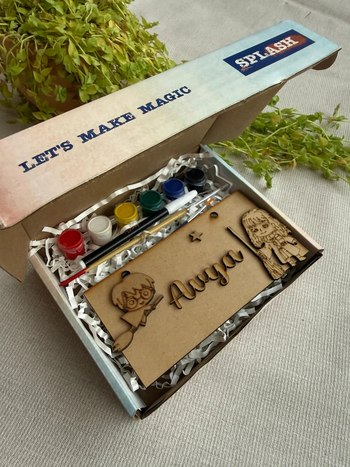 A personalized children's craft kit in an open cardboard box, featuring wooden cutout figures, acrylic paint bottles, a paintbrush, and a wooden nameplate engraved with 'Auya'. The box lid has 'SPLASH' branding and 'LET'S MAKE MAGIC' printed on it, set on a neutral fabric background with green foliage props.