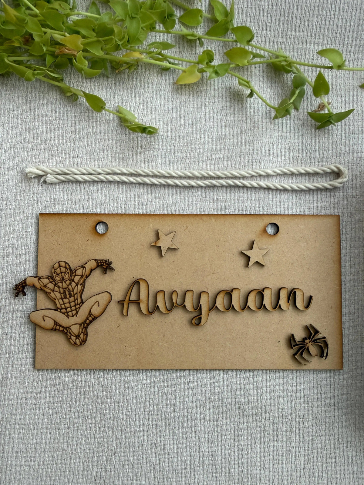 A personalized wooden nameplate engraved with the name 'Avyaan' and decorated with a laser-cut Spider-Man figure, stars, and a spider motif, shown on a textured light gray fabric background with a green plant sprig at the top.