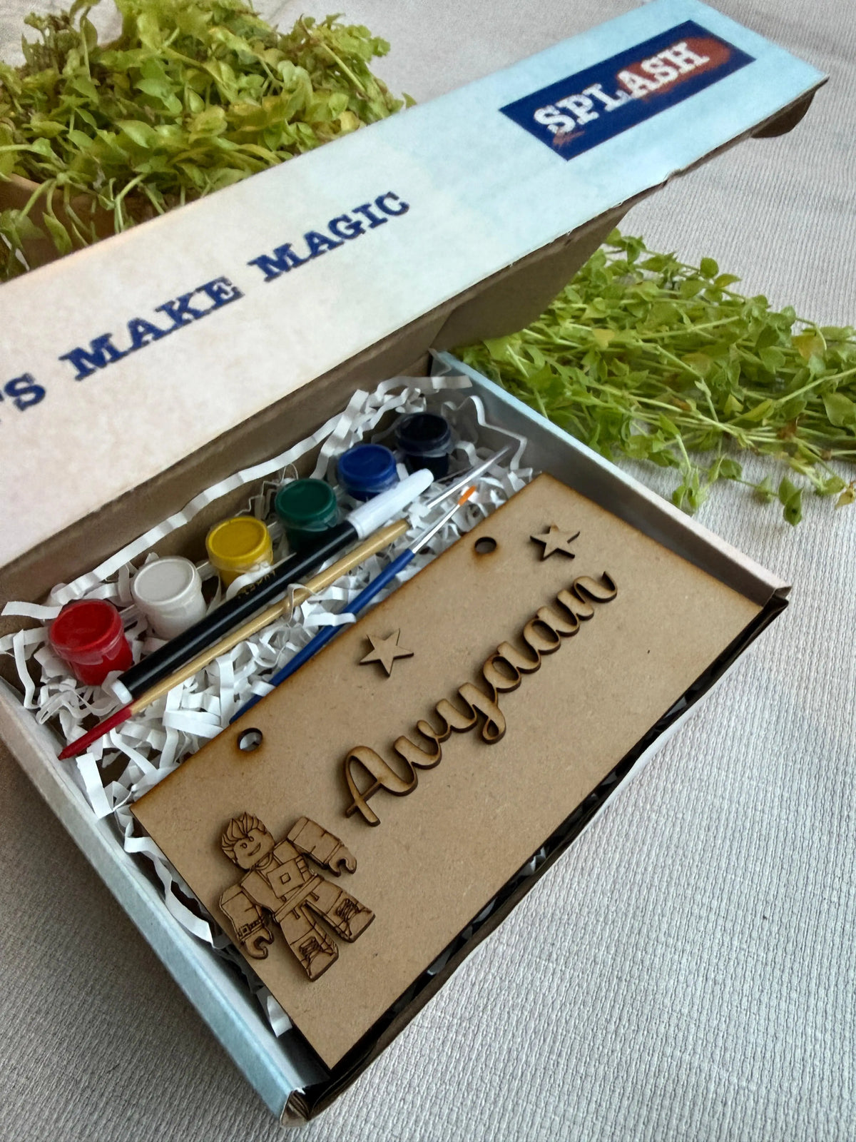 A personalized kids craft kit in a cardboard box, featuring acrylic paint pots, a paintbrush, and a wooden name plaque engraved with 'Aryaan' and decorated with laser-cut stars and a small robot design. The box lid has 'SPLASH' printed on it.