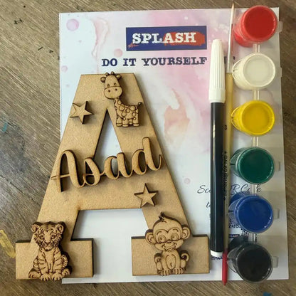 A DIY children's first birthday paint-by-numbers activity kit featuring a wooden personalized letter 'A' with animal cutouts and the name 'Asad'. The kit includes a paint palette with red, white, yellow, green, blue, and black paint cups, a paint brush, and an instruction sheet on a wooden table.