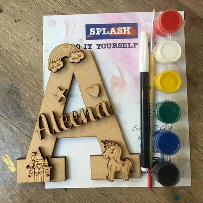 Personalized wooden letter A craft kit with the name 'Aleena', a rainbow, stars, a unicorn, and small illustrations, shown with a paint palette, paint brush, and marker on a wooden tabletop.
