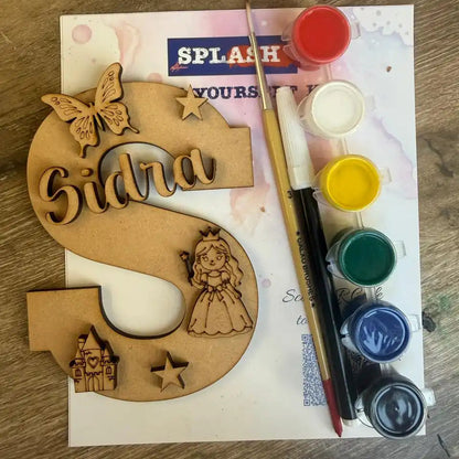 A personalized wooden letter 'S' craft kit with the name 'Sidra' laser-cut, decorated with a butterfly, star, princess silhouette, and castle, shown with a paintbrush and a set of colorful acrylic paints on a wooden table.