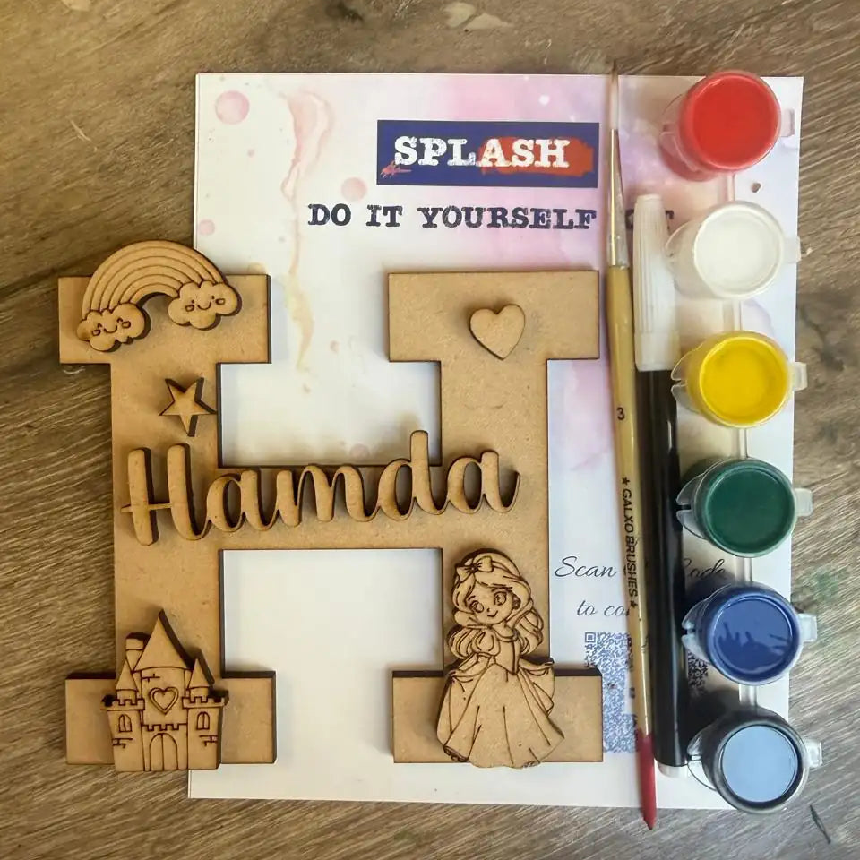 A wooden personalized letter H craft kit on a rustic wooden table, featuring laser-cut wooden figures spelling 'Hamda', a rainbow, a castle, and a princess design, along with a paint palette, paint pots, and a paintbrush.