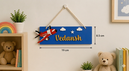 Decorative wall hanging with airplane and 'Vedansh' text, placed on a shelf with teddy bears and books.