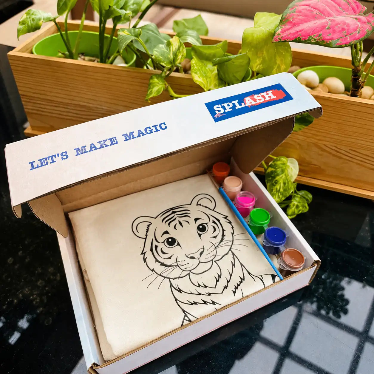 Box with a tiger drawing and paint supplies, labeled 'Splash', on a reflective surface with plants in the background.
