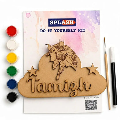 A DIY laser-cut and paint kit shown on a white background, including a printed card with a Batman illustration, a cloud-shaped wooden cutout with the name Tamizh, five paint pots, a fine-tip brush, and a marker pen.