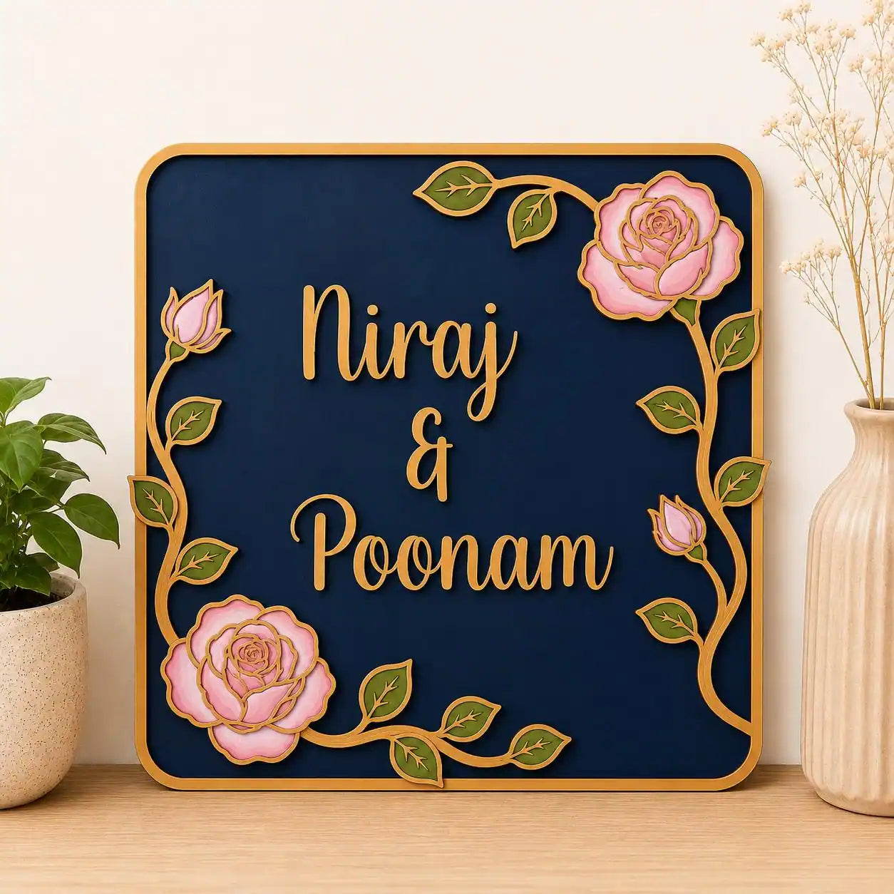 Decorative wall art with 'Niraj & Poonam' text surrounded by pink roses and green leaves on a navy background.