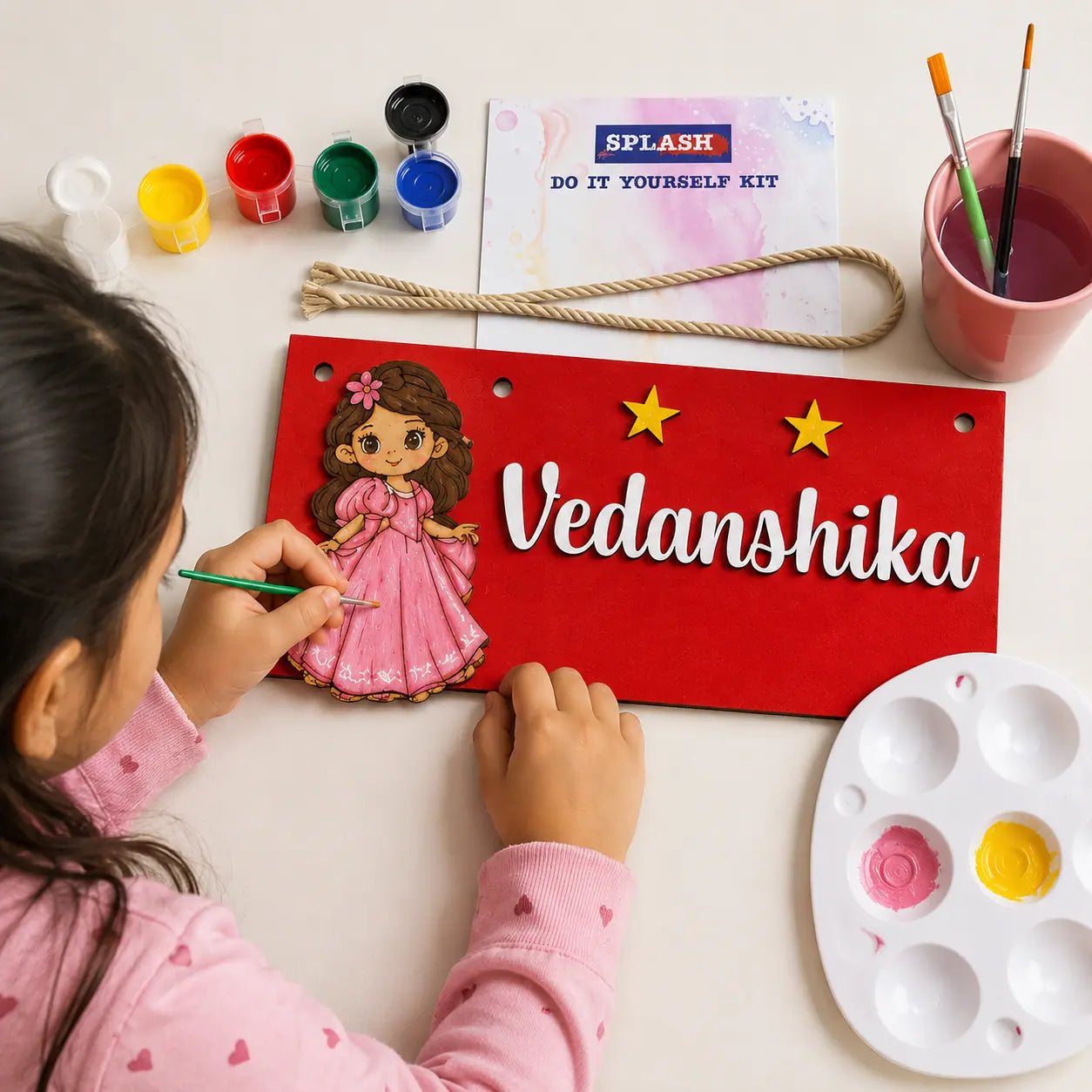 Child painting a nameplate with a doll illustration and 'Vedanshika' on a white table.