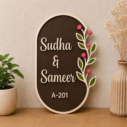 Decorative sign with 'Sudha & Sameer' and house number 'A-201' on a wooden frame with floral designs, placed on a surface with a plant and vase in the background.