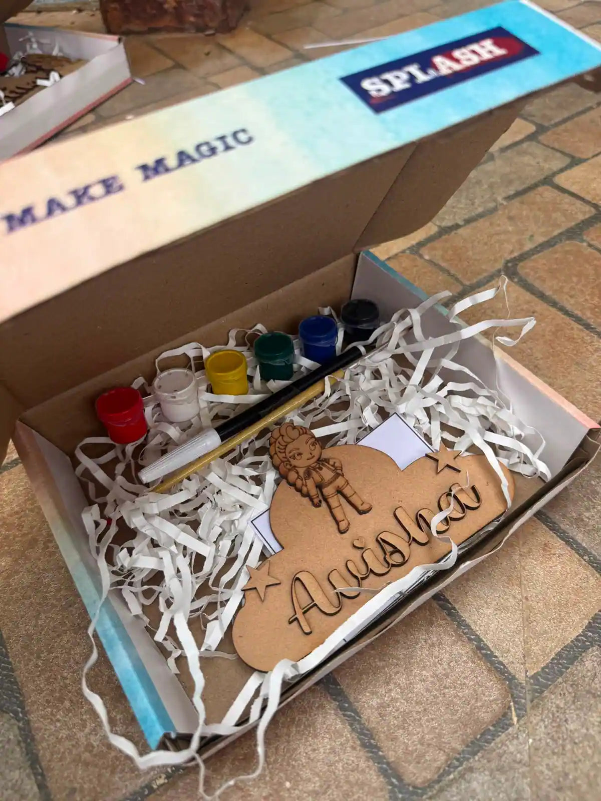 An open cardboard gift box filled with white paper shred contains a personalized wooden name sign reading 'Amishka' with a lion illustration, plus small paint pots, a paintbrush, and a marker. The box lid has 'SPLASH' printed on it.