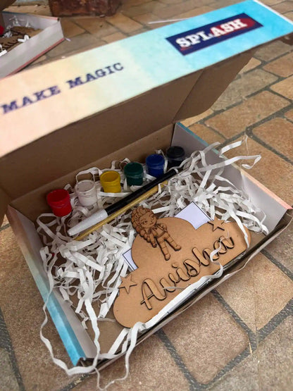An open cardboard gift box filled with white paper shred contains a personalized wooden name sign reading 'Amishka' with a lion illustration, plus small paint pots, a paintbrush, and a marker. The box lid has 'SPLASH' printed on it.