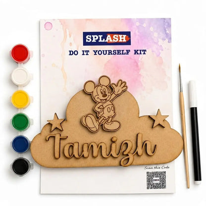 A DIY wooden name carving kit shown flat on a white background, with a printed instruction card reading SPLASH and DO IT YOURSELF KIT, a laser-cut wooden cloud plaque with a Mickey Mouse figure and the name Tamizh, paint pots on the left, a paintbrush, and a marker on the right.