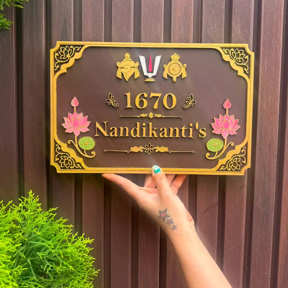 A rectangular custom wooden nameplate with a dark brown background and ornate gold border, held by a hand against a dark brown wooden fence. The plaque features gold lettering reading '1670 Nandikanti's' with pink lotus flowers and green lily pads, and a small medal emblem at the top.