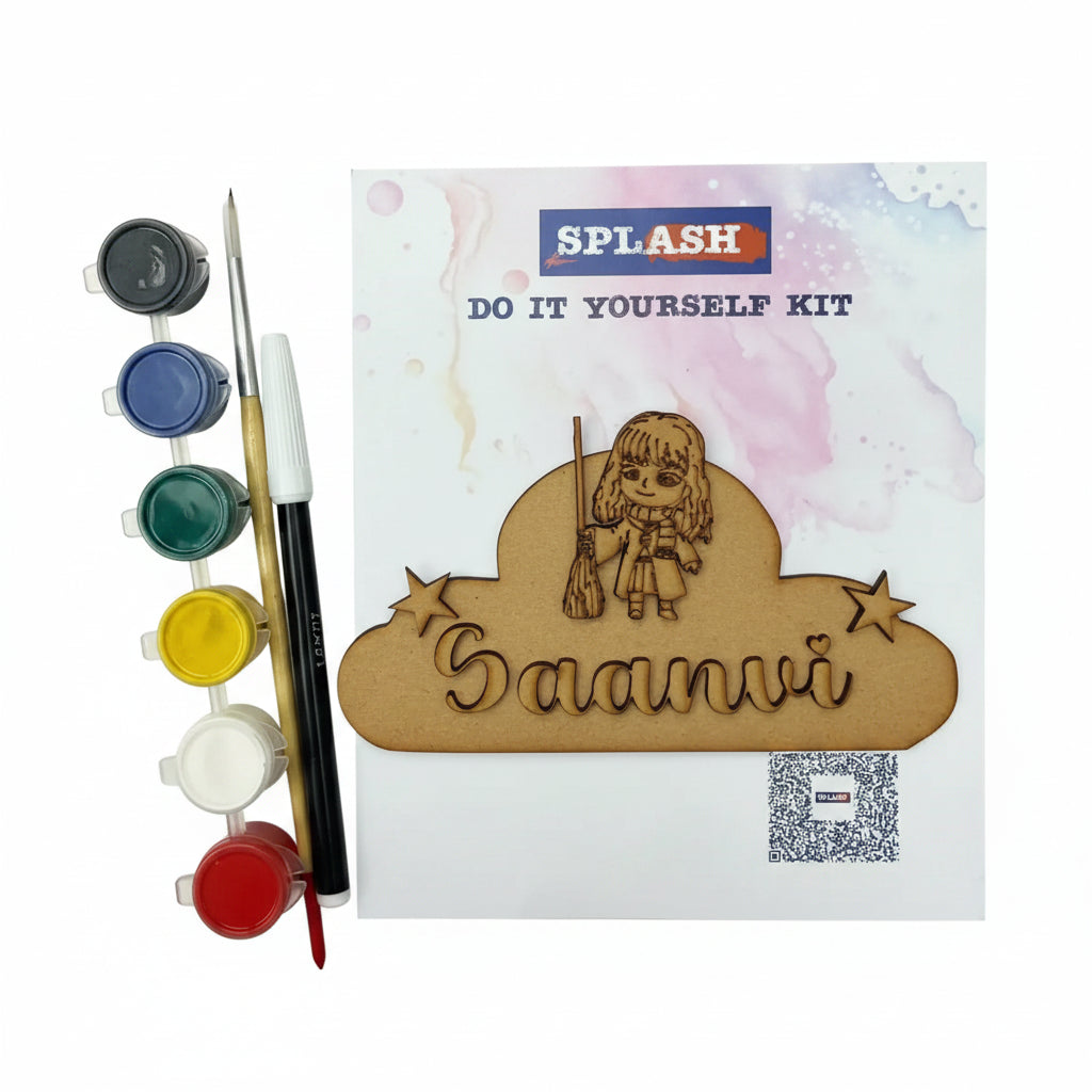 DIY craft kit labeled SPL.ASH DO IT YOURSELF KIT on a textured beige fabric background, with paint pots, a fine-tip pen, and a wooden nameplate featuring the engraved name 'Saanvi' and a cartoon girl holding a broom.