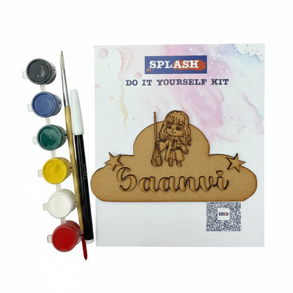 DIY craft kit labeled SPL.ASH DO IT YOURSELF KIT on a textured beige fabric background, with paint pots, a fine-tip pen, and a wooden nameplate featuring the engraved name 'Saanvi' and a cartoon girl holding a broom.